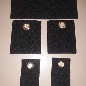 May include: Five black fabric squares with gold metal eyelets. The squares are different sizes and are arranged in a row.
