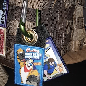 May include: A blue and white patch with the text "Baskin's Tiger Patch Challenge" and a cartoon tiger holding a box of cereal. The text "More-R-re Weight!" is below the tiger. The patch is attached to a brown bag with a green carabiner.