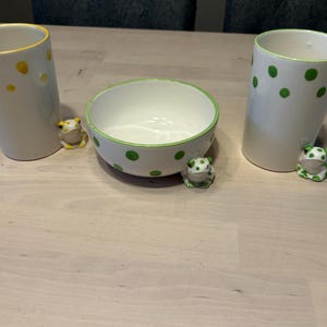 May include: Two white ceramic tumblers and one white ceramic bowl, each featuring hand-painted green or yellow polka dots and a small, sculpted frog figure attached to the base. The items have smooth, glossy finishes and simple, functional shapes.