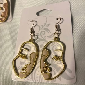 May include: A pair of gold-tone earrings with a white face design. The earrings are on a white card with a floral pattern.