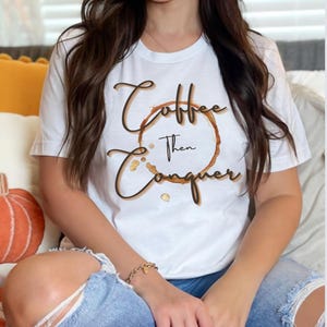 以下が含まれることがあります： 白いTシャツに、茶色のコーヒーのシミと、茶色の文字で「Coffee then Conquer」と書かれています。