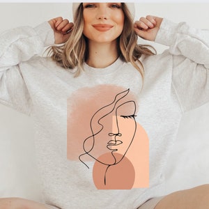 May include: A light gray sweatshirt with a graphic of a stylized face in black and peach tones. The face is outlined in black and has closed eyes and a slightly open mouth.