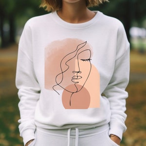 May include: White sweatshirt with a graphic design of a stylized face with closed eyes and a peach and pink watercolor background.