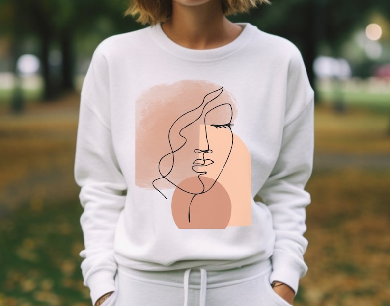May include: White sweatshirt with a graphic design of a stylized face with closed eyes and a peach and pink watercolor background.