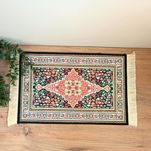 Pode incluir: Um tapete decorativo retangular com borda preta e franjas creme. O tapete apresenta um design central em forma de diamante em vermelho e creme, rodeado por um padr&atilde;o floral em v&aacute;rias cores.