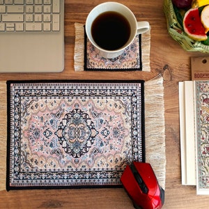 May include: A black and pink patterned mouse pad with fringed edges. The mouse pad is on a wooden surface with a laptop, a cup of coffee, a book, and a bookmark.