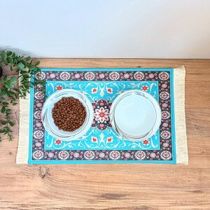 Peut inclure: Deux bols en c&eacute;ramique blanche avec un motif bleu et rouge sur un tapis turquoise &agrave; franges. Un bol est rempli de nourriture pour chien brune.