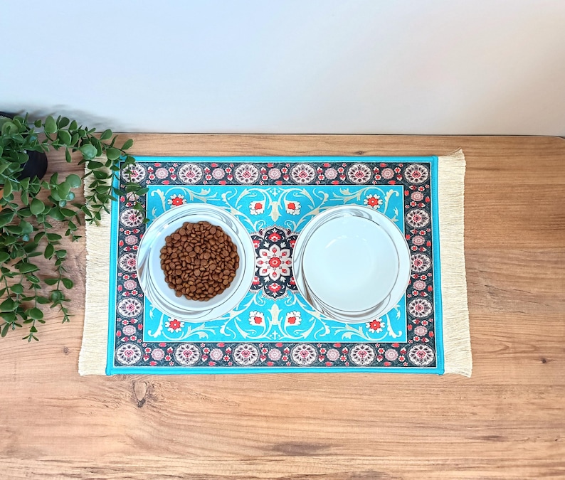 Peut inclure: Deux bols en c&eacute;ramique blanche avec un motif bleu et rouge sur un tapis turquoise &agrave; franges. Un bol est rempli de nourriture pour chien brune.