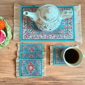 May include: Set of four turquoise blue coasters with intricate floral patterns in red, white, and gold. The coasters are made of fabric and have a fringed edge. A white teapot with a floral design sits on a larger turquoise blue coaster with a fringed edge.