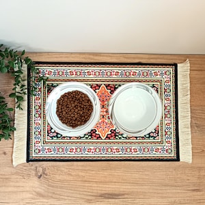 Pode incluir: Um tapete decorativo para alimenta&ccedil;&atilde;o de animais de estima&ccedil;&atilde;o com duas tigelas brancas. Uma tigela &eacute; preenchida com ra&ccedil;&atilde;o marrom, a outra est&aacute; vazia. O tapete tem um design colorido com bordas franjadas, assemelhando-se a um pequeno tapete.