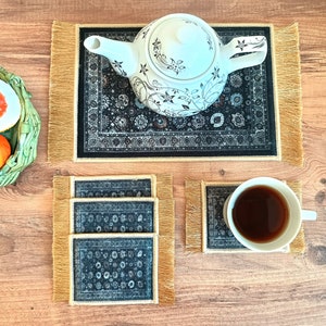 May include: A set of four black and gold coasters with a floral pattern. The coasters are made of fabric and have a fringe border. A white teapot with a floral design is sitting on a black and gold coaster with a fringe border.