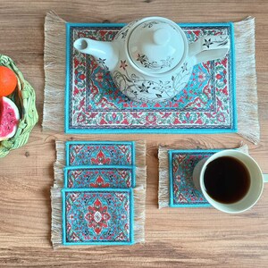 May include: Set of four turquoise blue and white patterned coasters with a floral design. The coasters are made of fabric and have fringe around the edges. A white teapot with a floral design sits on a matching coaster.