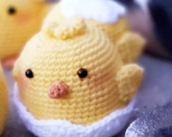 May include: A crocheted yellow chick with black eyes and a pink beak, sitting inside a white crocheted egg.