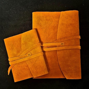 Può includere: Due diari in pelle scamosciata arancione con lacci in pelle. Il diario più grande ha tre lacci e il diario più piccolo ha due lacci.