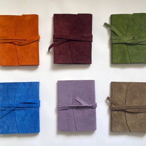 Könnte beinhalten: Sechs ledergebundene Notizbücher in verschiedenen Farben: Orange, Burgunder, Grün, Blau, Lila und Braun. Jedes Notizbuch hat ein schlichtes Design mit einem einzigen Riemenverschluss.