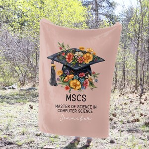May include: A pink fleece blanket with a black graduation cap decorated with colorful flowers. The text "MSCS Master of Science in Computer Science Jennifer" is printed on the blanket.