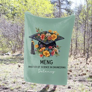 May include: A mint green blanket featuring a graduation cap design adorned with colorful flowers. The cap is black with a gold tassel. Text includes "MENG", "Master of Science in Engineering", and a name.