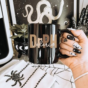 May include: Black coffee mug with the name "Valerie" written in gold lettering. The mug is being held by a hand with black and white nail polish featuring moons and stars.