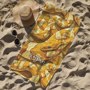 May include: A rectangular beach towel featuring a yellow and brown snake skin pattern with the name Logan printed in white script. A wide-brimmed straw hat with a white band and black sunglasses are positioned next to the towel on the sand.