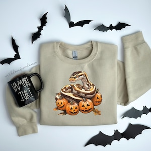 May include: A tan sweatshirt with a graphic of a snake curled around a pile of pumpkins. The snake is brown and tan with a light brown head. The pumpkins are orange with black jack-o-lantern faces. The sweatshirt has a crew neck and long sleeves.
