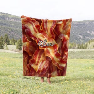 May include: A large, rectangular blanket featuring a realistic print of crispy bacon strips in shades of red and brown. The name "Alex" is written in cursive. The blanket is held up in a grassy field with a mountain backdrop.