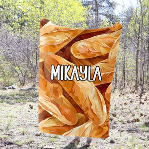 May include: A soft, brown and beige blanket with a pattern of baguette bread. The blanket has the name "Mikayla" printed on it in white.