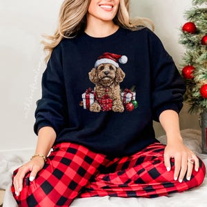 May include: A black sweatshirt with a Christmas-themed design featuring a brown and white dog wearing a Santa hat and surrounded by presents. The dog is sitting on a red and black plaid background.