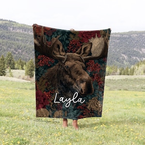 May include: A plush blanket featuring a detailed illustration of a moose with large antlers, set against a backdrop of dark floral patterns and red berries. The name "Layla" is written in cursive. The blanket is held outdoors in a field.