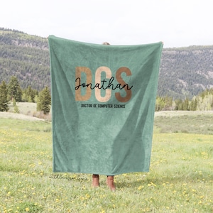 May include: A teal blanket with the text "DCS Jonathan DOCTOR OF COMPUTER SCIENCE" in brown and black lettering. The blanket is held up in a field with a mountain range in the background. The blanket is a soft, cozy item.