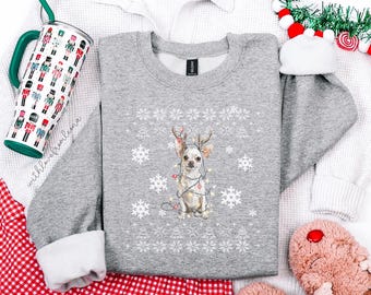 May include: A gray sweatshirt with a festive design featuring a Chihuahua wearing reindeer antlers and Christmas lights. The sweatshirt is surrounded by a white waffle-weave shirt, a tumbler with a nutcracker design, and red and white checkered fabric.