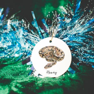 May include: A round white ceramic ornament featuring a detailed illustration of a coiled brown and tan ball python snake. The name Henry is printed in black script lettering below the snake. A gold string is attached to the top for hanging.