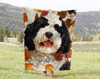 May include: A white and black dog with brown eyes and a pink tongue is featured on a fleece blanket. The blanket has a fall theme with orange, yellow, and red leaves. The dog's name, "Koda", is written in black cursive on the blanket.