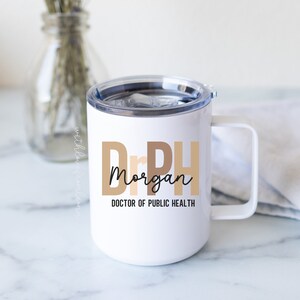 May include: White travel mug with a handle and a clear lid. The mug features the text "Dr PH Morgan DOCTOR OF PUBLIC HEALTH" in a mix of fonts and colors. The mug is on a white surface.
