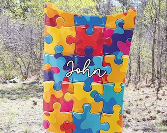 May include: A colorful fleece blanket with a puzzle piece pattern in shades of yellow, blue, red, and pink. The name "John" is written in white cursive on the blanket.