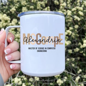 May include: White stainless steel travel mug with a black handle. The mug has a gold and brown design with the text "McCoupe Alexandria" and "Master of Science in Computer Engineering". The bottom of the mug has the text "with love from leona".