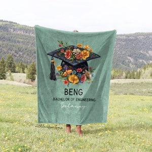 May include: A teal blanket featuring a graduation cap design adorned with colorful flowers. The cap has a black tassel and is surrounded by orange, yellow, and pink flowers. The blanket has the text "BENG Bachelor of Engineering Delaney".
