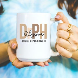 May include: A white ceramic mug with the text "Dr. PH Alyssa DOCTOR OF PUBLIC HEALTH" in brown and black lettering. The mug is held by a person with multiple gold rings on their fingers.