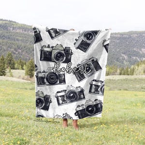 May include: White fleece blanket with a repeating pattern of vintage cameras in black and white. The name "Robert" is written in cursive across the blanket.