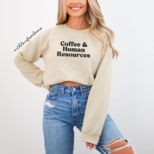 May include: A beige long-sleeve crewneck sweatshirt featuring the black text Coffee & Human Resources printed on the chest. The garment has a ribbed neckline, cuffs, and hem, and is paired with blue denim jeans that have distressed detailing.