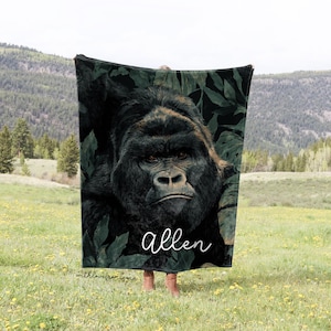 May include: A dark green blanket featuring a detailed image of a gorilla's face surrounded by dark green leaves. The name "Allen" is written in white script. The blanket is held up in a field with mountains in the background.