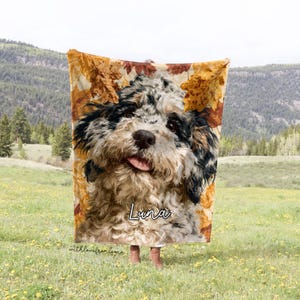 May include: A rectangular fleece blanket featuring a large, detailed portrait of a black and white curly-haired dog. The background of the blanket is decorated with golden-brown autumn leaves. The name Luna is printed in white script text.