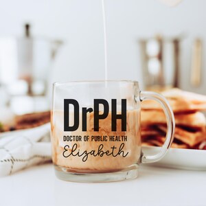 May include: A clear glass mug with a handle, filled with a light brown liquid. The mug has the text "DrPH DOCTOR OF PUBLIC HEALTH Elizabeth" in black. Cream is being poured into the mug. The mug is on a white surface.