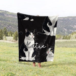 May include: Black blanket featuring a repeating pattern of a black and white cat with green eyes. The name "Katie" is written in cursive. The blanket is held up in a field with mountains in the background.