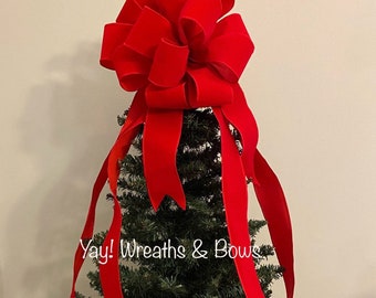May include: A red velvet Christmas tree topper bow with long tails. The bow is tied around a small potted Christmas tree. The text "Yay! Wreaths & Bows" is visible on the tree.