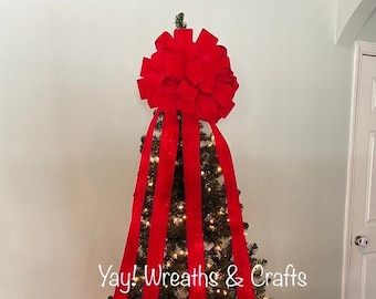 May include: A red velvet Christmas tree topper bow with four long streamers. The bow is on top of a green artificial Christmas tree with white lights. The tree is in a wicker basket. The text "Yay! Wreaths & Crafts" is visible on the tree.