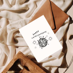 Pode incluir: Um cartão branco com as palavras "happy cultivarsary" e uma ilustração de bola de discoteca. O cartão é acompanhado por um envelope castanho e repousa sobre um tecido creme. Estrelas decorativas rodeiam a bola de discoteca.
