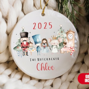 May include: A white ceramic Christmas ornament featuring a Nutcracker theme. The ornament has the year 2025 in red, along with illustrations of Nutcracker characters, a clock, and a Christmas tree. The name "Chloe" is also displayed.
