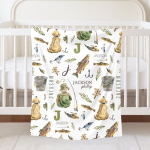 May include: A white baby blanket with a fishing theme. The blanket features a repeating pattern of fish, fishing rods, lures, a bucket, a dog, and the letter "J". The text "JACKSON phillip" is also included on the blanket.