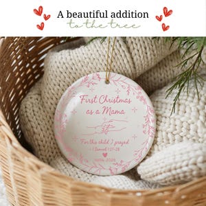 May include: A white ceramic Christmas ornament with pink floral accents and the text "First Christmas as a Mama." The ornament includes the text "For this child I prayed - 1 Samuel 1:27-28" and the name "Hallie 2026."