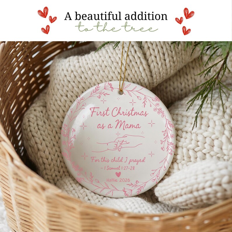 May include: A white ceramic Christmas ornament with pink floral accents and the text "First Christmas as a Mama." The ornament includes the text "For this child I prayed - 1 Samuel 1:27-28" and the name "Hallie 2026."
